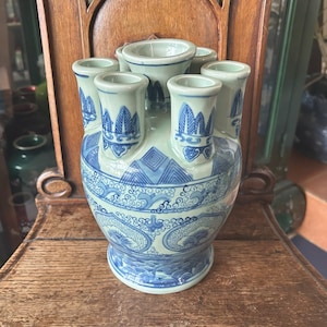 May include: A blue and white ceramic tulip vase with multiple openings for flowers. The vase features a detailed pattern of stylized waves, mountains, and floral motifs. The vase is sitting on a wooden surface.