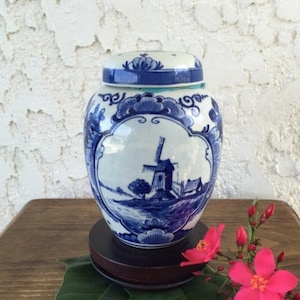 May include: A blue and white ceramic jar with a lid, featuring a windmill scene. The jar sits on a dark wooden base, with pink flowers and green leaves in the foreground. The jar is approximately 6 inches tall.