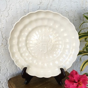 Puede incluir: Un plato de cer&aacute;mica blanco con un borde festoneado y un dise&ntilde;o en relieve en el centro. El plato est&aacute; sobre un soporte de madera.