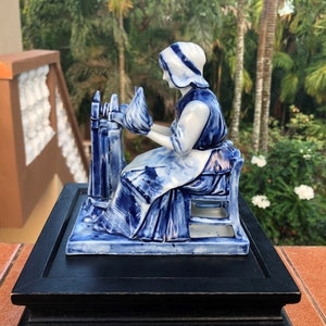 K&ouml;nnte beinhalten: Eine blau-wei&szlig;e Porzellanfigur einer Frau, die Garn spinnt. Die Figur sitzt, tr&auml;gt eine Haube und eine Sch&uuml;rze und arbeitet an einem Spinnrad. Die Figur ist auf einem schwarzen Holzsockel montiert.