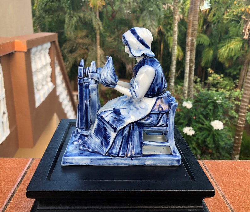 K&ouml;nnte beinhalten: Eine blau-wei&szlig;e Porzellanfigur einer Frau, die Garn spinnt. Die Figur sitzt, tr&auml;gt eine Haube und eine Sch&uuml;rze und arbeitet an einem Spinnrad. Die Figur ist auf einem schwarzen Holzsockel montiert.