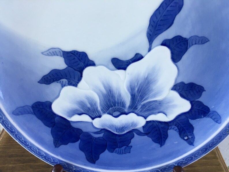 Op de afbeelding: Een wit en blauw keramisch bord met een bloemmotief. Het bord heeft een grote, gestileerde witte bloem met blauwe accenten en bladeren. De rand heeft een decoratief blauw patroon.