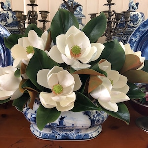 Op de afbeelding: Een bloemstuk met grote witte magnoliabloemen met groene en bruine bladeren. De bloemen zijn gerangschikt in een blauw-witte keramische vaas met een Hollandse molen scène. Decoratieve items zijn op de achtergrond.