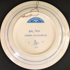 May include: Back of a white ceramic plate with blue concentric lines and text. The text includes "De Porceleyne Fles", "216/500", and "DESIGN: JTH:DESSENS". A copper wire is attached for hanging.
