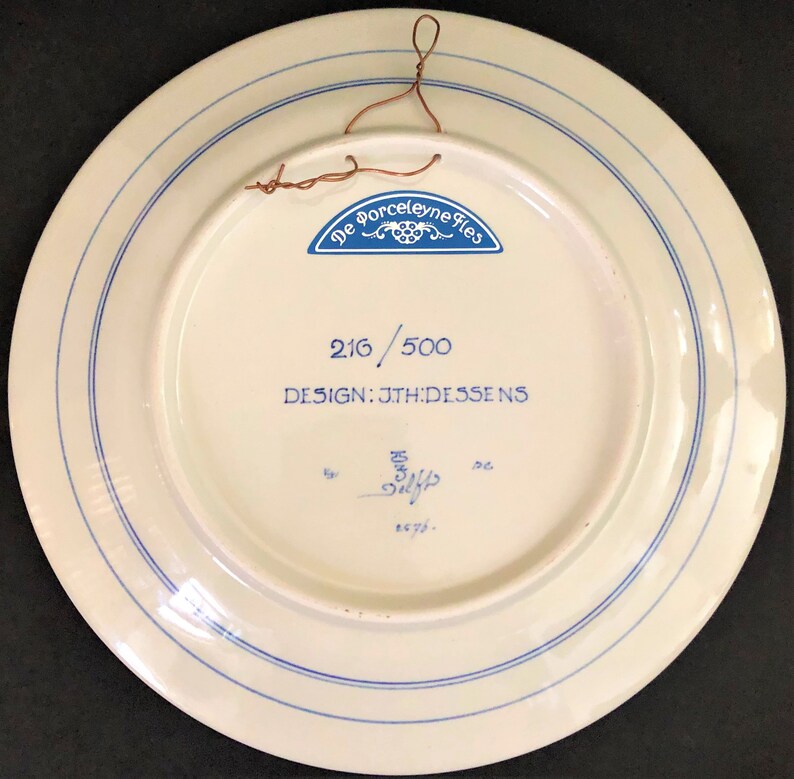 May include: Back of a white ceramic plate with blue concentric lines and text. The text includes "De Porceleyne Fles", "216/500", and "DESIGN: JTH:DESSENS". A copper wire is attached for hanging.