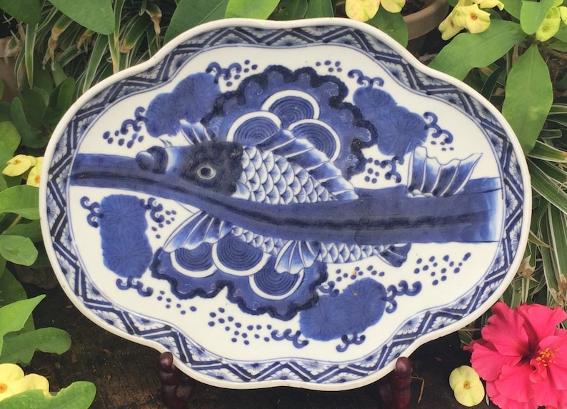 K&ouml;nnte beinhalten: Ein wei&szlig;-blauer Keramikteller mit Fischmuster. Der Teller hat einen gewellten Rand mit einem geometrischen Rand. Der Fisch ist in Blau dargestellt, mit detaillierten Schuppen und Flossen. Der Teller dient wahrscheinlich dekorativen oder Servierzwecken.