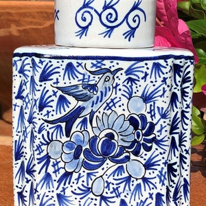 Puede incluir: Un recipiente de cer&aacute;mica blanca con tapa a juego, ambos adornados con dise&ntilde;os florales y de p&aacute;jaros azules. El recipiente es rectangular con bordes redondeados. La tapa presenta un patr&oacute;n de remolino. El dise&ntilde;o recuerda a la cer&aacute;mica de Delft.