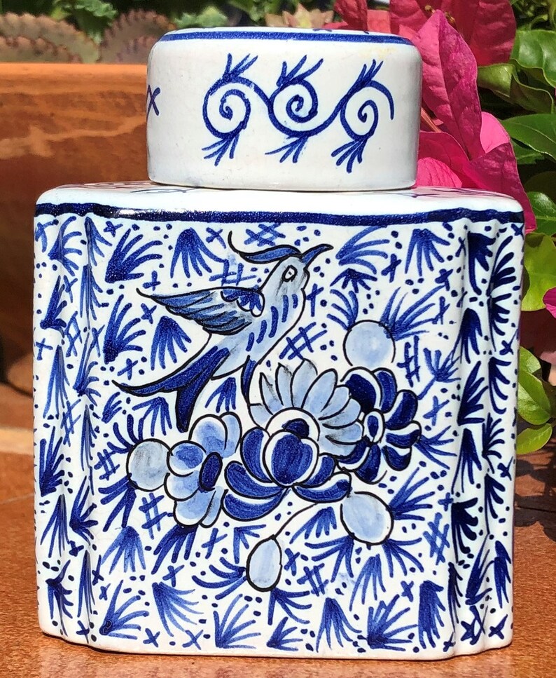 Puede incluir: Un recipiente de cer&aacute;mica blanca con tapa a juego, ambos adornados con dise&ntilde;os florales y de p&aacute;jaros azules. El recipiente es rectangular con bordes redondeados. La tapa presenta un patr&oacute;n de remolino. El dise&ntilde;o recuerda a la cer&aacute;mica de Delft.
