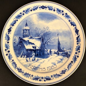 May include: A blue and white Delftware plate depicting a winter scene. The plate features a church, a house, and a canal with a sailboat. The plate is decorated with a holly border and the text "KERSTMIS 1983".