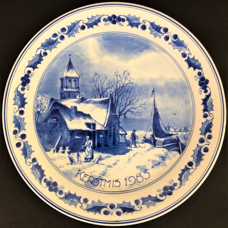 May include: A blue and white Delftware plate depicting a winter scene. The plate features a church, a house, and a canal with a sailboat. The plate is decorated with a holly border and the text "KERSTMIS 1983".