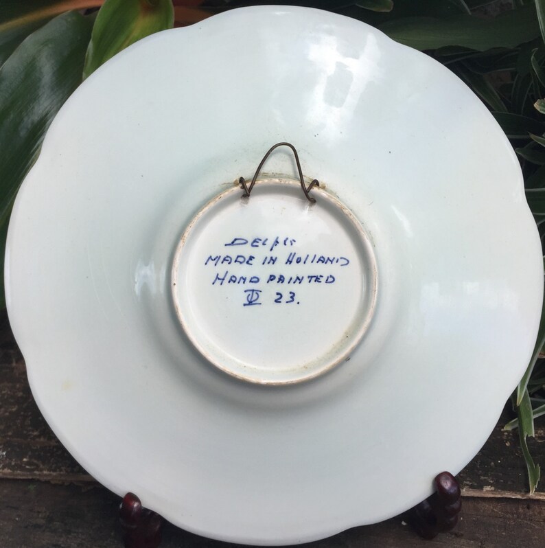 May include: A white ceramic plate with a blue and white hand-painted design. The back of the plate has the text "Delft Made in Holland Hand Painted D 23."