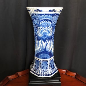 May include: A striking blue and white ceramic vase with a flared top and intricate floral patterns. The vase is displayed on a black stand. The design features detailed artwork, with a predominantly blue and white colour scheme, creating a classic look.