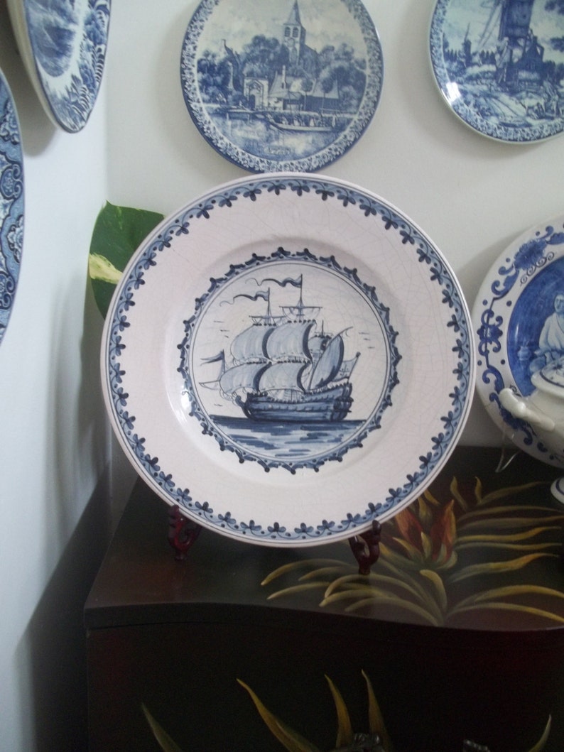 May include: A collection of blue and white decorative plates. The central plate features a detailed illustration of a sailing ship. Other plates depict scenes of buildings and landscapes. The plates are displayed on a dark wooden surface.