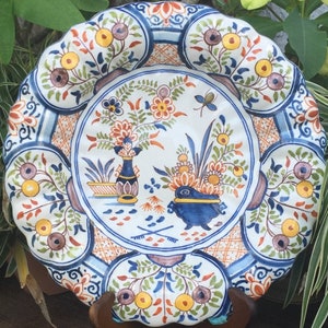 K&ouml;nnte beinhalten: Ein wei&szlig;er Keramikteller mit einem blauen und wei&szlig;en Blumendesign. Der Teller hat einen gewellten Rand und ist mit verschiedenen Blumen, Bl&auml;ttern und einem kleinen Schmetterling verziert.