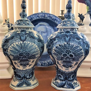 May include: Two matching blue and white ceramic vases with lids, featuring intricate floral and foliate patterns. The vases have a bulbous shape and are decorated with a detailed, hand-painted design. A matching plate is visible in the background.