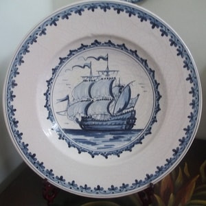 May include: White ceramic plate with a blue ship illustration in the center. The plate has a blue floral border and a decorative blue trim around the ship. The plate is likely for decorative purposes.