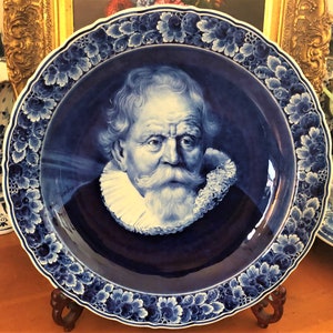 Puede incluir: Un plato de Delftware azul y blanco con un retrato de un hombre con un cuello de encaje blanco y una barba blanca. El plato tiene un borde floral azul y blanco.