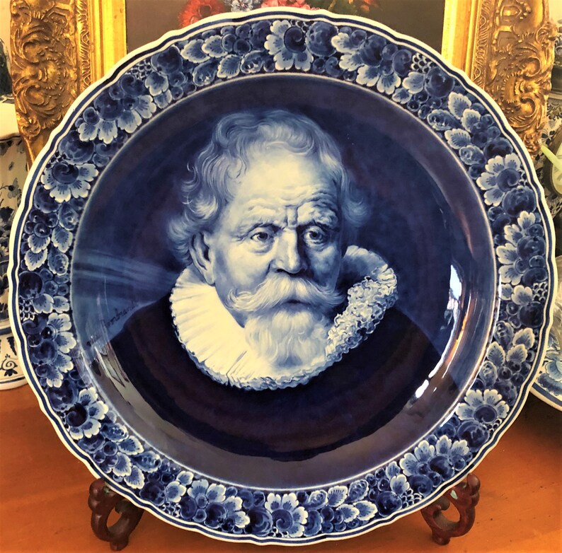 Puede incluir: Un plato de Delftware azul y blanco con un retrato de un hombre con un cuello de encaje blanco y una barba blanca. El plato tiene un borde floral azul y blanco.