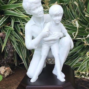 Puede incluir: Una escultura de porcelana blanca que representa a una persona arrodillada abrazando a un ni&ntilde;o. Las figuras est&aacute;n sobre una base octogonal marr&oacute;n oscuro. La escultura est&aacute; situada sobre un fondo de follaje verde y blanco.
