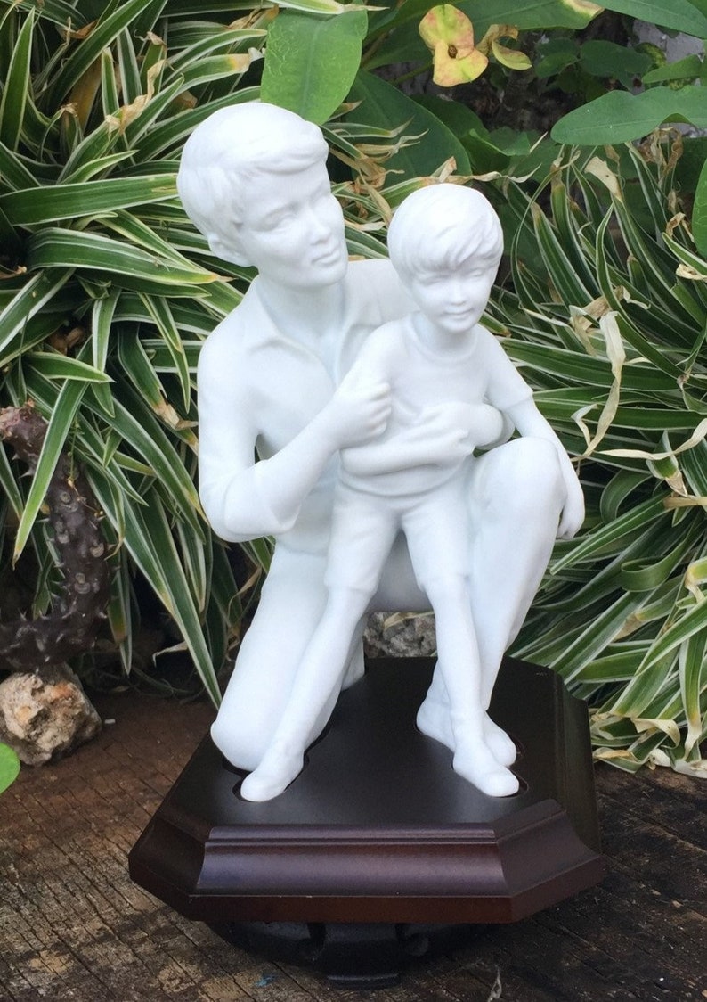 Puede incluir: Una escultura de porcelana blanca que representa a una persona arrodillada abrazando a un ni&ntilde;o. Las figuras est&aacute;n sobre una base octogonal marr&oacute;n oscuro. La escultura est&aacute; situada sobre un fondo de follaje verde y blanco.