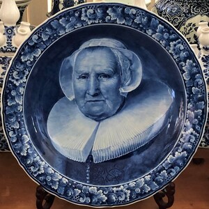 K&ouml;nnte beinhalten: Ein blauer und wei&szlig;er Teller aus Delft mit einem Portr&auml;t einer Frau, die einen wei&szlig;en Spitzenkragen und eine wei&szlig;e Haube tr&auml;gt. Der Teller ist mit einem floralen Rand verziert.