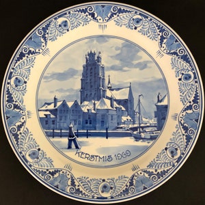 May include: A blue and white Delftware plate featuring a winter scene of a church and buildings. The plate has a blue and white floral border and the text "KERSTMIS 1969".