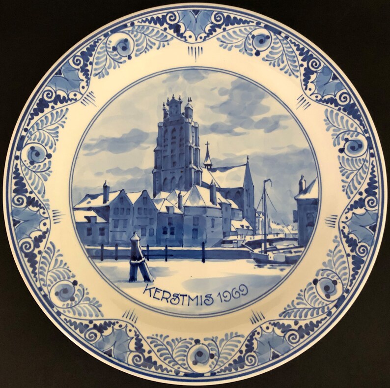 May include: A blue and white Delftware plate featuring a winter scene of a church and buildings. The plate has a blue and white floral border and the text "KERSTMIS 1969".