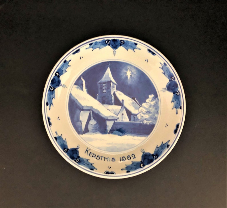 May include: A blue and white ceramic plate featuring a winter scene of a church and houses. The plate has a blue border with a holly leaf design and the text "Kerstmis 1962".