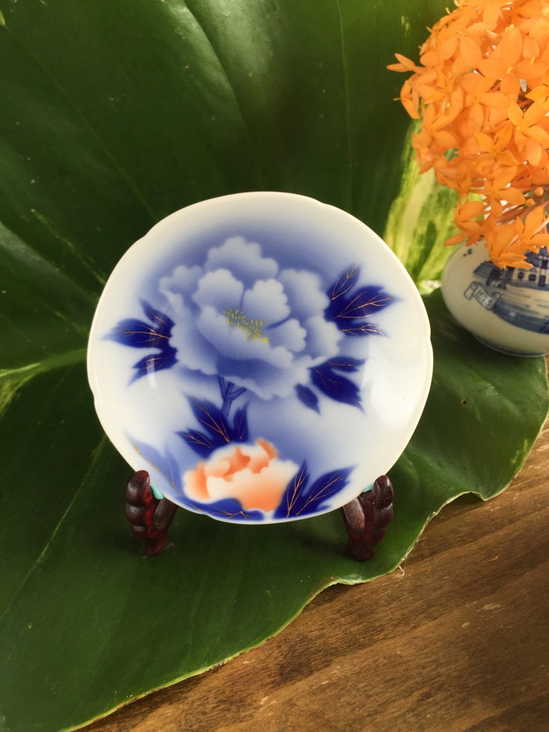 May include: A white and blue porcelain dish with a floral design, on a small wooden stand. The dish features a blue peony with gold accents and an orange flower. The background includes a large green leaf and orange flowers in a vase.