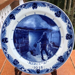 May include: A white and blue ceramic plate with a winter scene. The plate features a central image of a person and a dog near a building, with the word "KERSTMIS" and the year "1918" below. The rim has a blue floral design.
