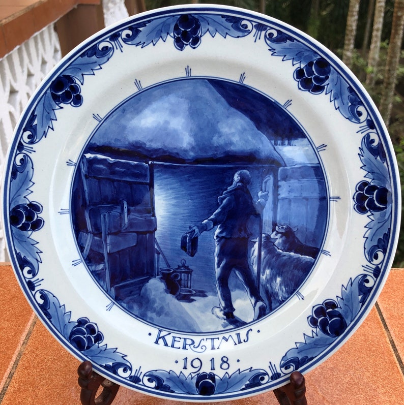 May include: A white and blue ceramic plate with a winter scene. The plate features a central image of a person and a dog near a building, with the word "KERSTMIS" and the year "1918" below. The rim has a blue floral design.