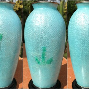 Pu&ograve; includere: Un vaso in ceramica blu chiaro con una superficie testurizzata e disegni floreali verdi. Il vaso &egrave; su un piedistallo nero.