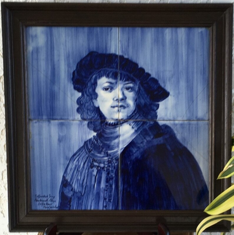 May include: A blue and white ceramic tile depicting a portrait of a person wearing a dark blue hat and a blue and white patterned jacket. The tile is framed in a dark wood frame.