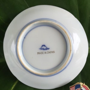 May include: A small, white ceramic plate with a blue rim and a blue mountain and text that reads "MADE IN JAPAN". A pin with the American and Dutch flags is next to the plate.
