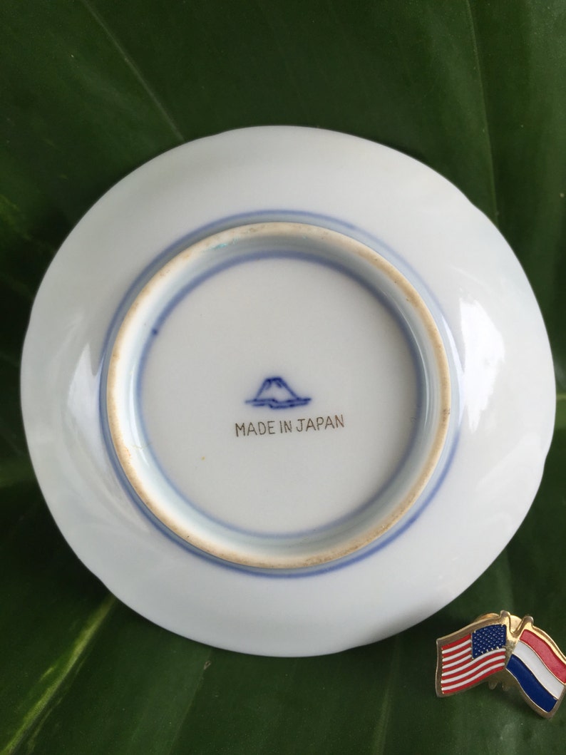 May include: A small, white ceramic plate with a blue rim and a blue mountain and text that reads "MADE IN JAPAN". A pin with the American and Dutch flags is next to the plate.