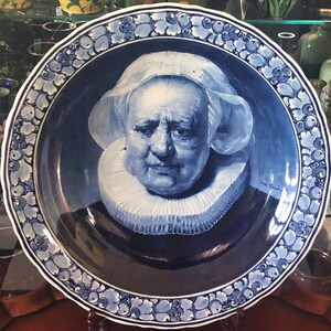 May include: A blue and white Delftware plate featuring a portrait of a woman wearing a white lace collar and a white cap. The plate has a decorative border of blue and white floral designs.