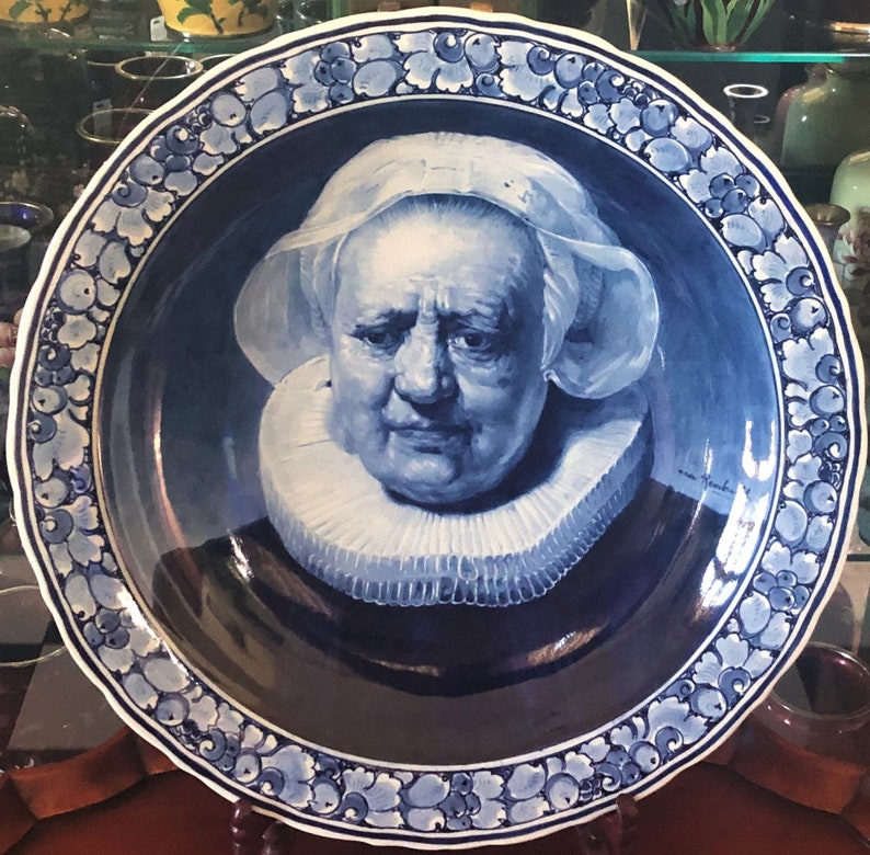 May include: A blue and white Delftware plate featuring a portrait of a woman wearing a white lace collar and a white cap. The plate has a decorative border of blue and white floral designs.