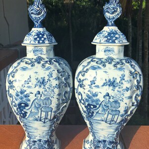 May include: Two matching blue and white ceramic vases with lids. The vases feature intricate floral and landscape designs, with a figure seated near a table. The lids are topped with a flame-like finial. The vases are displayed on a brown surface.