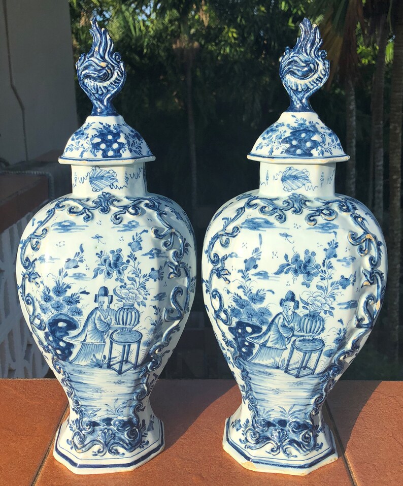 May include: Two matching blue and white ceramic vases with lids. The vases feature intricate floral and landscape designs, with a figure seated near a table. The lids are topped with a flame-like finial. The vases are displayed on a brown surface.