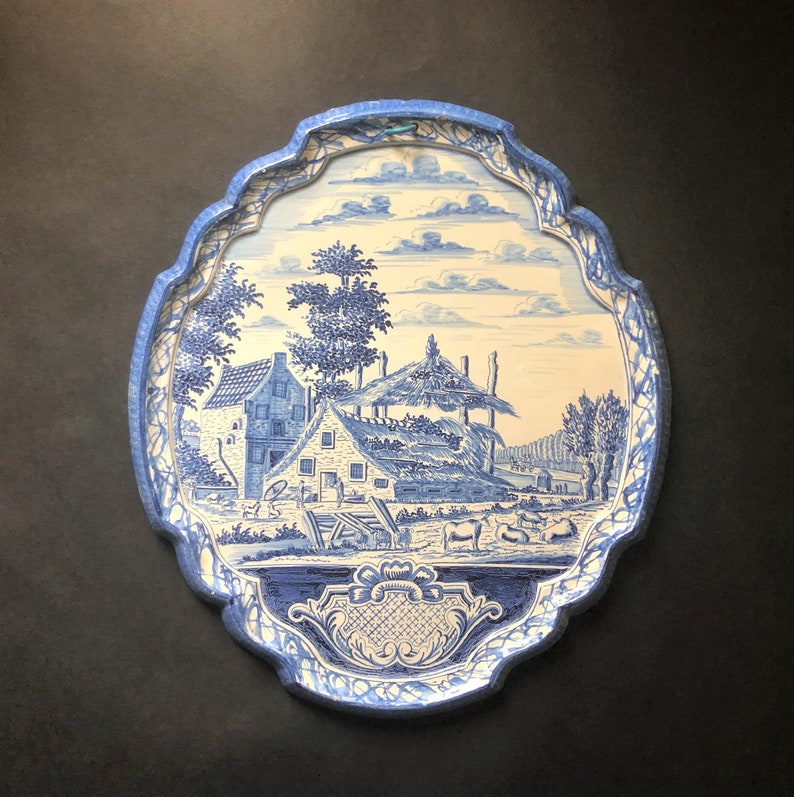 May include: Oval-shaped blue and white decorative tray with a pastoral scene. The tray features a detailed illustration of a village with houses, trees, and livestock. The border is a scalloped design in a matching blue.