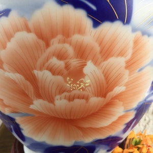 Puede incluir: Primer plano de un jarr&oacute;n de cer&aacute;mica con un gran dise&ntilde;o de peon&iacute;a de color melocot&oacute;n. La flor est&aacute; centrada, rodeada de patrones florales azules y blancos con detalles dorados. El jarr&oacute;n tiene un acabado brillante.