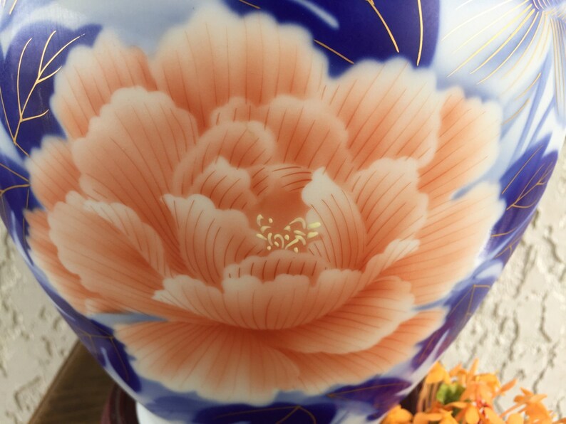 Puede incluir: Primer plano de un jarr&oacute;n de cer&aacute;mica con un gran dise&ntilde;o de peon&iacute;a de color melocot&oacute;n. La flor est&aacute; centrada, rodeada de patrones florales azules y blancos con detalles dorados. El jarr&oacute;n tiene un acabado brillante.