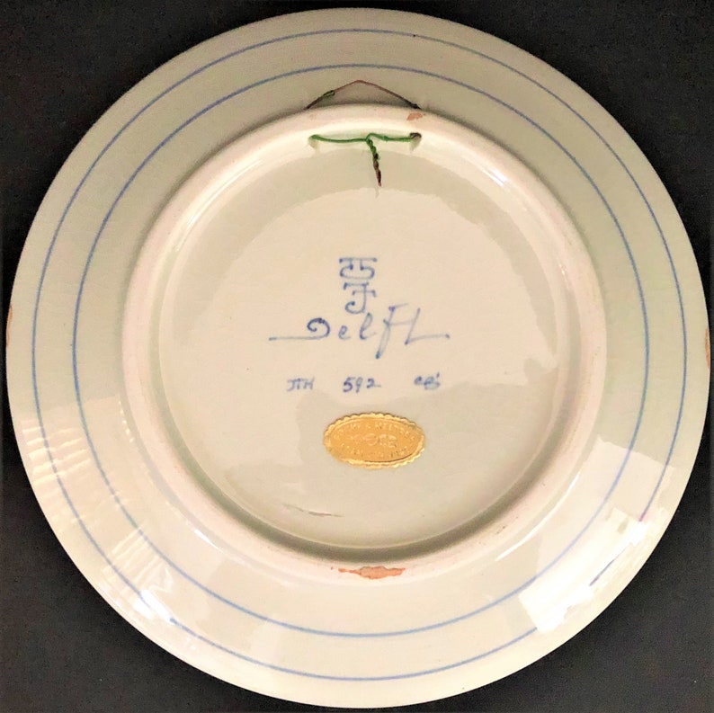 May include: A white ceramic plate with blue and green accents. The back of the plate has a gold sticker with the text "DeLft" and the number "592".