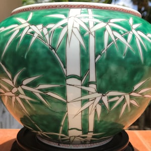 Puede incluir: Un jarrón redondo de cerámica verde con un diseño de bambú. El jarrón presenta tallos y hojas de bambú blancos sobre un fondo verde. El borde tiene un patrón rojo y blanco. El jarrón se asienta sobre una base negra.