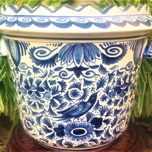 May include: A white and blue ceramic planter with a scalloped rim and two handles. The planter is decorated with a detailed blue floral and bird pattern. The planter is sitting on a wooden stand.