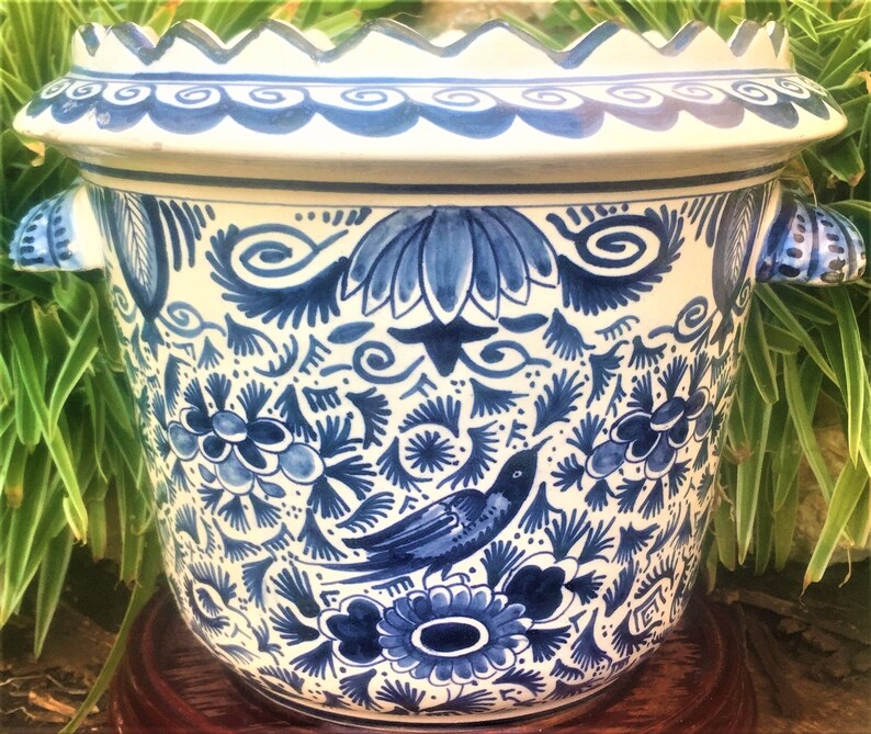 May include: A white and blue ceramic planter with a scalloped rim and two handles. The planter is decorated with a detailed blue floral and bird pattern. The planter is sitting on a wooden stand.