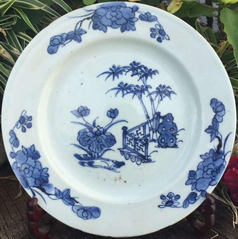 Op de afbeelding: Een wit porseleinen bord met een blauw en wit bloemenontwerp. Het bord heeft een centraal ontwerp van een bamboebos met een hek en een rots. De rand van het bord is versierd met een doorlopend bloemenpatroon.