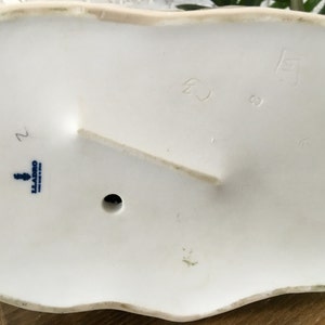 Puede incluir: Parte inferior de una base de figura de porcelana blanca con un borde ondulado. La base tiene un peque&ntilde;o agujero y un soporte elevado. Texto azul y un logotipo est&aacute;n estampados en la base. Tambi&eacute;n son visibles n&uacute;meros y marcas.