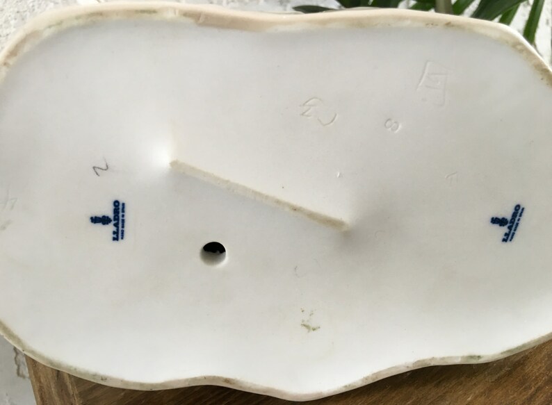 Puede incluir: Parte inferior de una base de figura de porcelana blanca con un borde ondulado. La base tiene un peque&ntilde;o agujero y un soporte elevado. Texto azul y un logotipo est&aacute;n estampados en la base. Tambi&eacute;n son visibles n&uacute;meros y marcas.