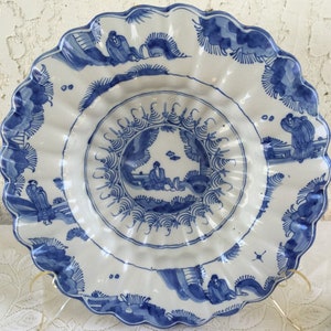 K&ouml;nnte beinhalten: Ein gro&szlig;er, wei&szlig;er Teller aus Keramik mit einem blauen und wei&szlig;en Design. Der Teller hat einen gewellten Rand und zeigt ein zentrales Design einer Figur in einer Landschaft. Der Teller ist mit stilisierten B&auml;umen und Blumen verziert.